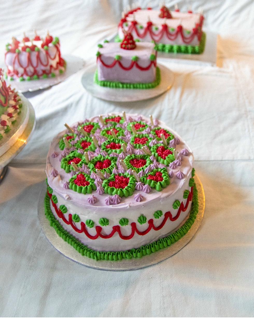 Celebration cake — red cherry topped cake with lavender, green and red piped details, multiple cakes displayed on linen