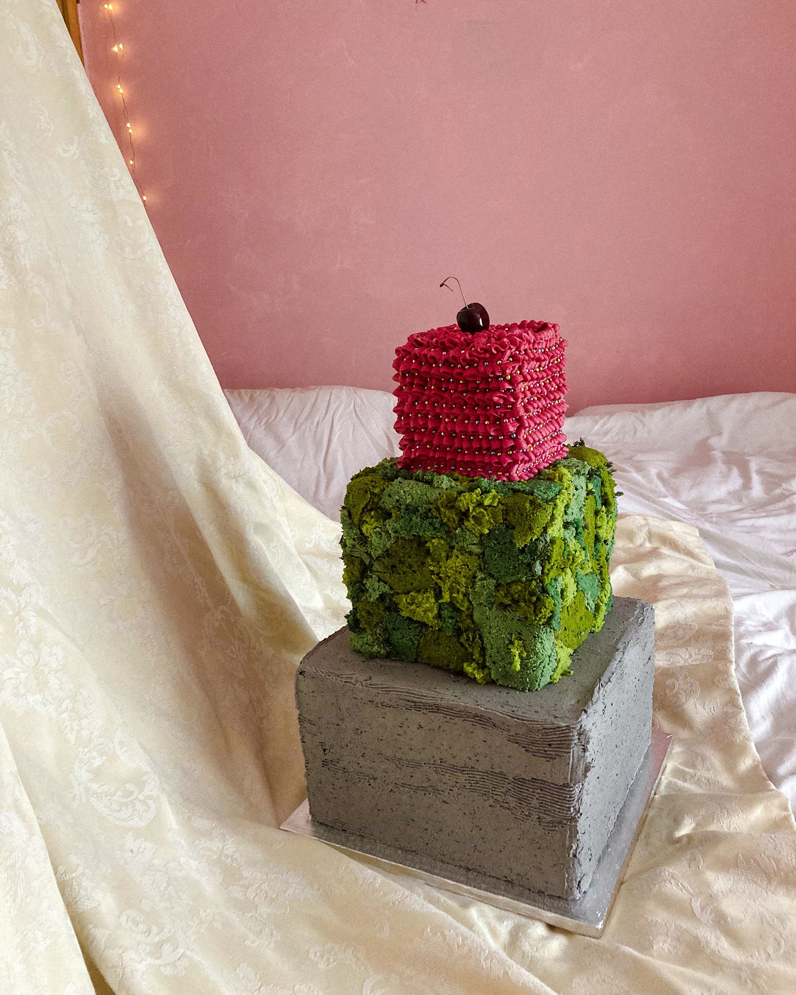 Celebration cake — three-tier sculptural cake with grey concrete-effect base, green moss-textured middle and pink ruffled piping top with cherry