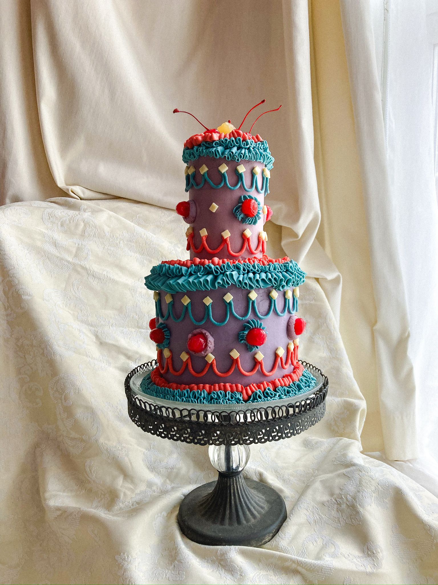 Celebration cake — two-tier mauve buttercream with teal and red piped ruffles, scallops, white chocolate squares and maraschino cherries on vintage glass stand